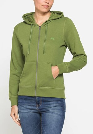 Donna che indossa una felpa verde con zip, tasche frontali e cappuccio, abbinata a jeans blu, su uno sfondo chiaro uniforme.