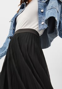 Kvinna i vitt linne, distressed denimjacka och flödande svart plisserad kjol mot en neutral bakgrund.