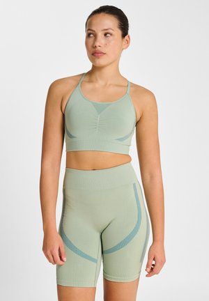 Vrouw die een lichtgroene naadloze sportbeha en bijpassende biker shorts met hoge taille en blauwe accenten draagt, staand tegen een effen achtergrond.