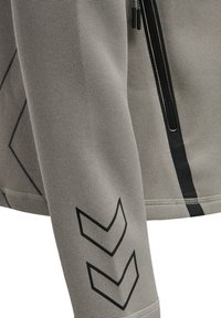 Gros plan d’une manche de veste grise avec un motif double chevron noir et une fermeture éclair noire verticale sur un tissu texturé.