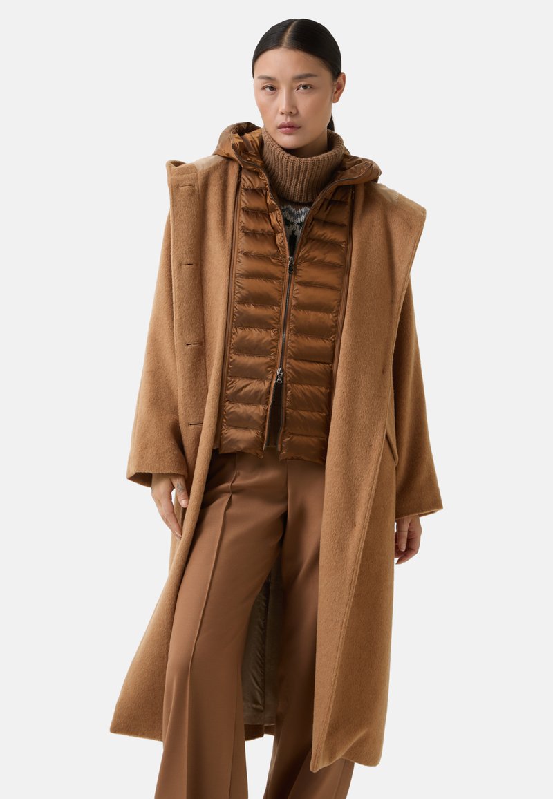 Brun uld frakke med hætte, lagd over en brun isoleret lynlåsvest og brune bukser, med en struktureret turtleneck under.