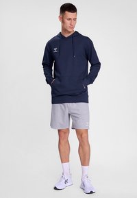 Marineblå hettegenser med snorer og kengurulomme, kombinert med lysegrå shorts. Modellen har på seg hvite treningssko og ankelsokker.