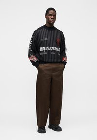 Pull rayé noir avec texte graphique, détail d'épaule orné, associé à un pantalon large marron et des baskets noires.