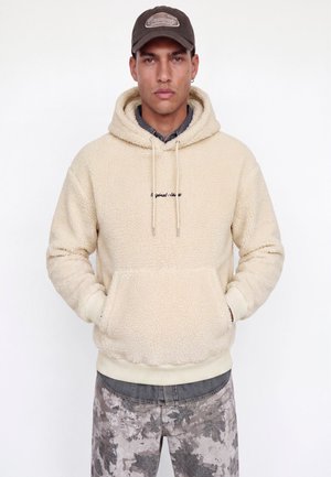 Homme portant un sweat à capuche sherpa beige avec poche avant, pantalon camouflage gris, chemise gris foncé en dessous et casquette de baseball marron.