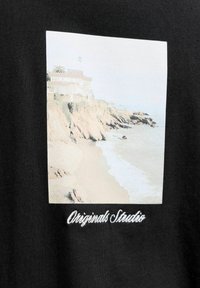 Strand bij een klif met golven, rotsachtige kustlijn en huizen bovenop, geprojecteerd op zwart textiel met de tekst "Originals Studio" eronder.