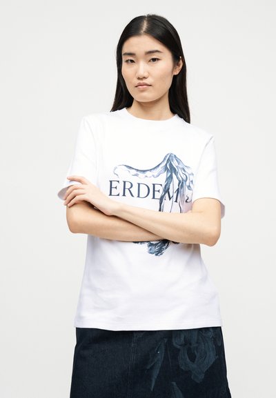 Νεαρή γυναίκα με μακριά μαύρα μαλλιά, φορώντας λευκό μπλουζάκι με γραφικό με χταπόδι και σκούρο φούστα, στέκεται με σταυρωμένα τα χέρια.