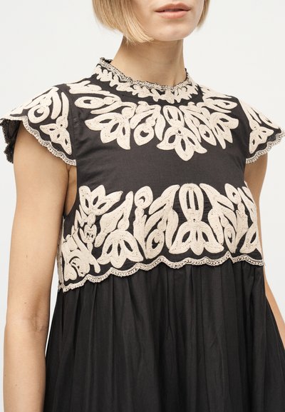 Γυναίκα που φοράει μαύρο αμάνικο φόρεμα με μπεζ ανθώδη κέντημα στο στήθος και στα μανίκια, με ψηλό λαιμόκοψη και κυματιστά τελειώματα.