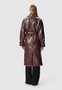 Person facing away wearing a long, shiny dark burgundy belted trench coat over black wide-leg pants.