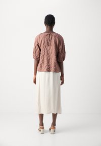 Braunes, strukturiertes Oberteil mit Puffärmeln und einem Rückenknoten, kombiniert mit einem weißen, fließenden, knielangen Rock und hellen, blockabsatz Schuhen.
