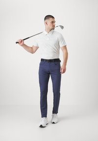 Bijela polo majica, navy hlače, crni pojas i bijele patike. Drži golf štap, stojeći naslonjen na jednostavnu bijelu pozadinu.