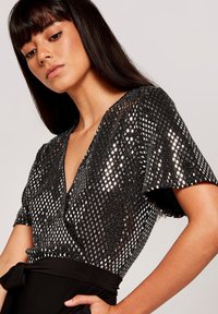 Top negro con escote en V y mangas cortas, con un patrón metálico texturizado que presenta formas de diamante. La tela suave contrasta con los lentejuelas.