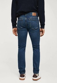 Jean bleu en denim avec une coupe slim, cinq poches et une pièce en cuir marron à l'arrière de la taille. Stylisé avec des baskets blanches et marron.