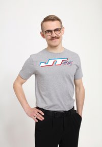 Grå bomulds t-shirt med et farverigt "JT Racing" logo i blå, rød og hvid. Modellen har sorte bukser og et sort bælte på.