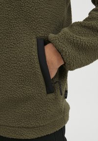 Fleece jacket in olive green with a textured surface; features black accents on the side pockets and a small logo tag.