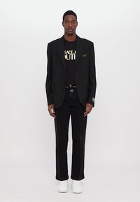 Blazer nero con rever a scatto su una t-shirt nera con testo dorato, abbinato a jeans neri e sneakers bianche; design minimalista.