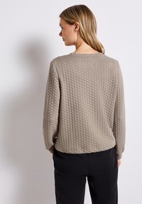 Texturerad beige tröja med diamantmönster, rund halsringning och långa ärmar, tillsammans med svarta byxor. Vy från baksidan.