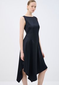 Robe noire sans manches avec une texture lisse et satinée, ourlet asymétrique et détails de couture en biais. Accentuée par de simples boucles d'oreilles.