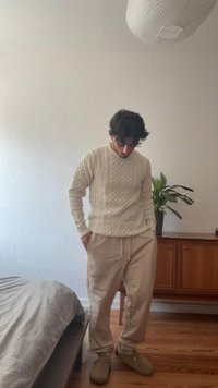 Krémový svetr s pleteným vzorem a béžové tepláky, s jemnou texturou. Osoba stojí vedle dřevěné skříně a květiny v květináči.