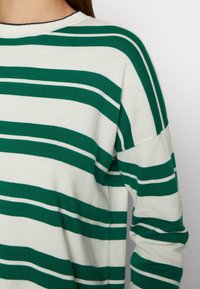Pull en tricot blanc avec de larges et étroites rayures horizontales vertes, montrant le haut du dos et le bras d'une personne.