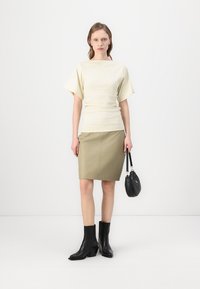 Haut crème avec des manches larges, jupe crayon verte olive, bottines noires à talons, et un petit sac à main noir. Lignes simples et textures lisses.
