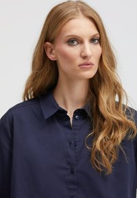 Navy button-up shirt with a collared neck, soft fabric, and visible button details, worn by a model with long wavy hair.