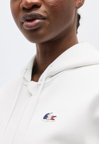 Lähikuva henkilöstä, joka käyttää valkoista vetoketjullista hupparia, jossa on sinivalkoverisen krokotiililogon vasemmassa rinnassa, yksiväristä taustaa vasten.