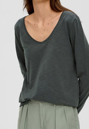 Woman wearing dark gray long-sleeve V-neck shirt and light gray pleated trousers against plain background.
