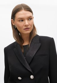 Czarna marynarka z teksturowanym czarnym kołnierzem, podwójnymi guzikami — jednym perłowym i jednym metalowym — oraz gładkim wykończeniem z delikatnymi prążkami.