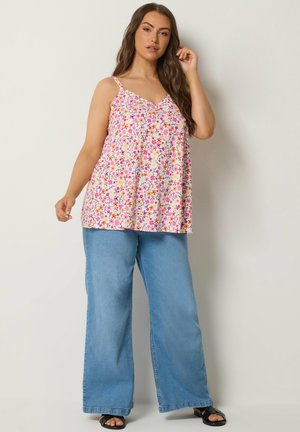 Kvinde iført en blomsterprikket ærmeløs top og vidde blå jeans, poserer med en hånd nær ansigtet mod en ensfarvet baggrund.