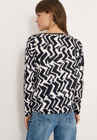 Langærmet top i bomuldsblanding med sort-hvidt abstrakt zigzag-mønster, rund halsudskæring og løs pasform. Kombineret med blå jeans.