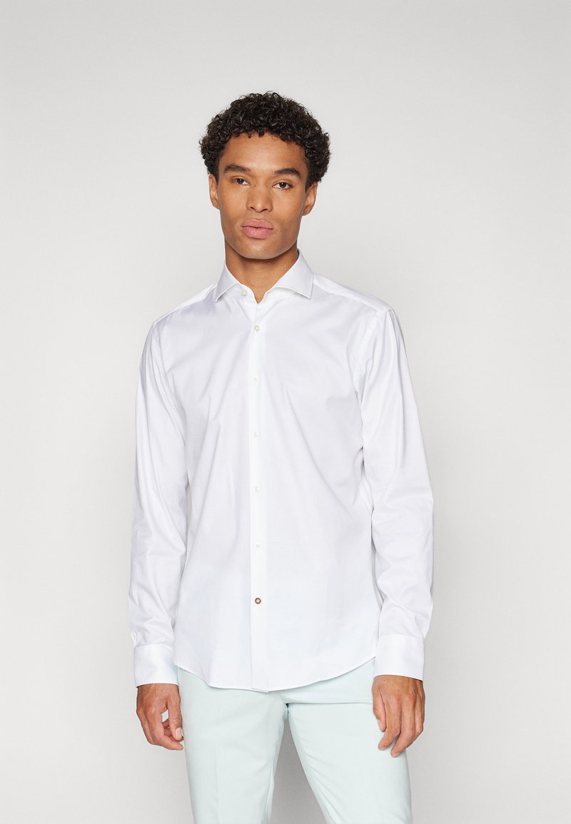 Camisa blanca de manga larga y botón, con un collar clásico, fabricada en tejido suave, con un ligero brillo y un solo botón decorativo en el cuello.