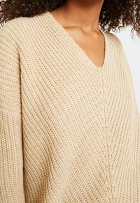 Beige pletený svetr s V-výstřihem, horizontální rib žebrovou strukturou a spadlými ramenními švy pro uvolněný střih.