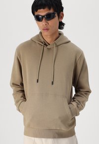 Beige hoodie with a front pocket, adjustable drawstring hood, ribbed cuffs, and hem. Model wears black sunglasses. Smooth texture.