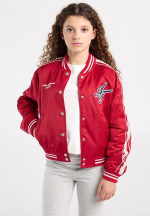 Jong meisje met krullend haar, gekleed in een rode varsityjas, wit shirt en lichtgrijze broek, staand tegen een effen achtergrond.