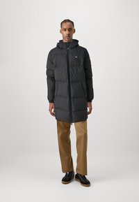 Zwarte lange pufferjas met een capuchon, doorgestikte ontwerp, ritssluiting aan de voorkant en elastische manchetten; gecombineerd met tan broek en zwarte schoenen.