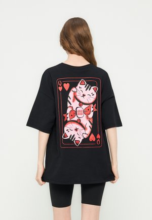 Woman with long brown hair wearing a black oversized T-shirt featuring a red and pink playing card design with two cats and "Good Luck" text.