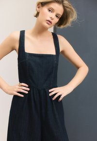 Dunkelblauer Jeans-Overall mit quadratischem Ausschnitt, breiten Trägern und geraffter Taille. Verfügt über ein schlichtes Design und eine glatte Textur.