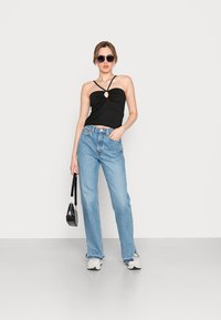 Top preto sem mangas com detalhe em nó e abertura, combinado com jeans retos de tom azul claro e ténis brancos. Pequena mala preta.