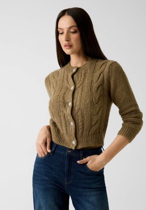 Donna con capelli lunghi e scuri che indossa un cardigan marrone a trecce con bottoni decorativi e jeans blu scuro, posando con le mani nelle tasche.