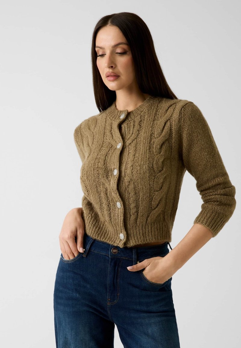 Femme aux longs cheveux bruns portant un cardigan marron à torsades avec des boutons décoratifs et un jean bleu foncé, posant les mains dans les poches.