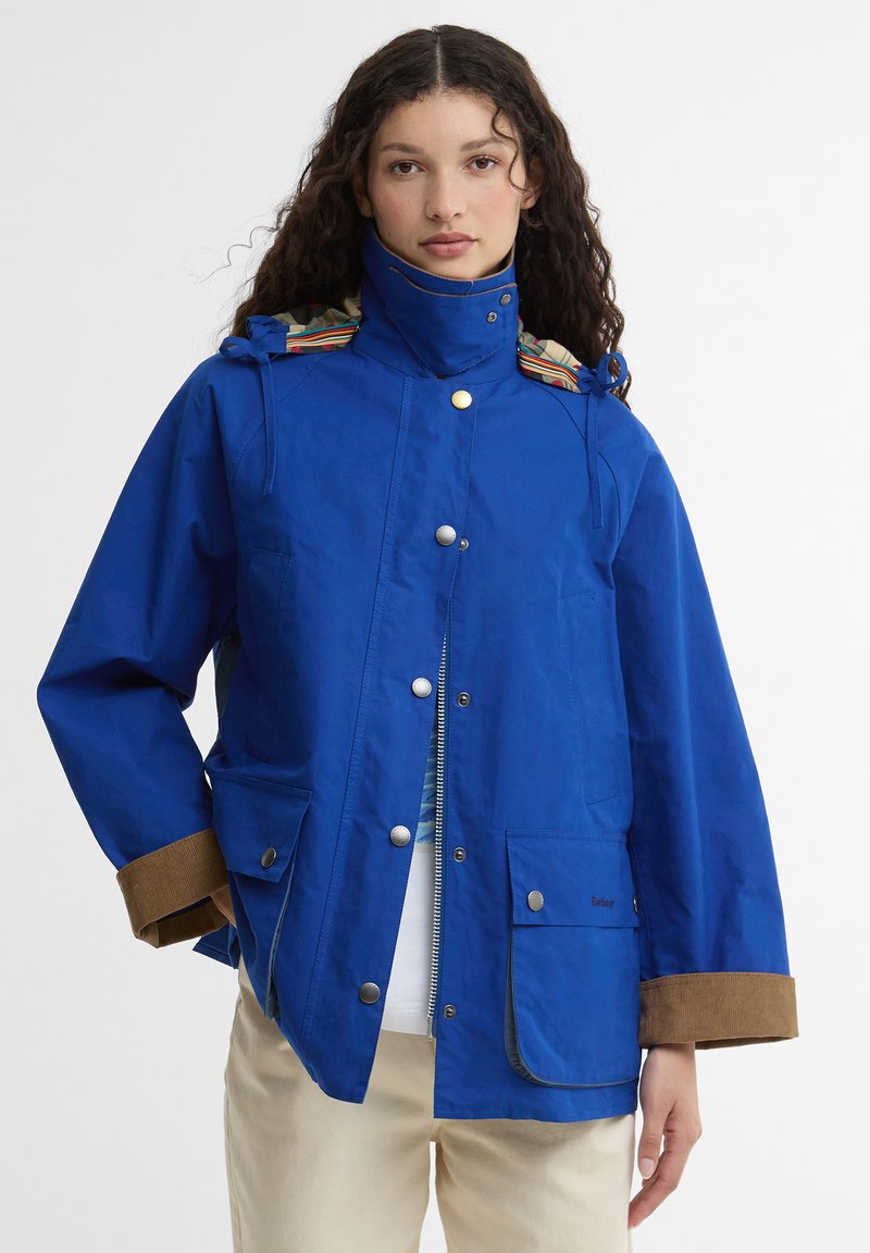 Γυναίκα με μακριά σγουρά μαλλιά που φορεί ένα μπλε παλτό βασιλικού τόνου με καφέ μανσέτες, κουμπιά και φερμουάρ μπροστά, όρθια μπροστά σε απλό φόντο.