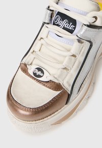 Bafalo sporta apavi ar krēmkrāsas augšdaļu, metālisku bronzas akcentu, sudraba detaļām, teksturētu zoli un melnu logo uz mēles.