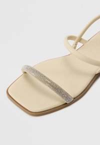 Sandal beige à lanières avec un bout carré, présentant une lanière ornée de strass argentés. Semelle lisse avec des éléments de design minimalistes.