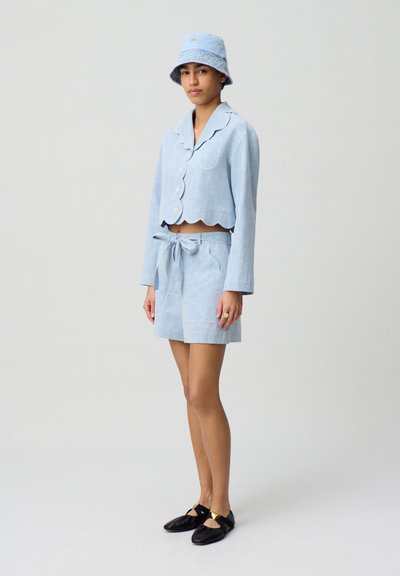 Chaqueta corta de manga corta en azul claro con bordes ondulados, combinada con pantalones cortos de tiro alto a juego y un sombrero de cubo, todo en una tela similar.