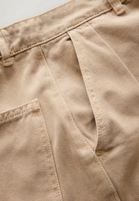 Close-up of beige cotton pants showing belt loops, front pocket, and detailed stitching on fabric.