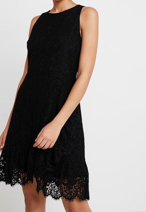 Ærmeløs sort knælange kjole i blonder med rund halsudskæring og bølget kant, båret af en person med let bøjede arme.