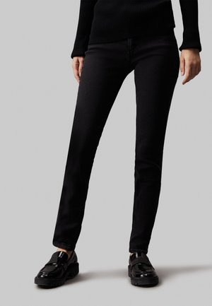 Jeans de mezclilla negra con un corte ajustado, costuras mínimas y una textura suave. Combinados con mocasines negros brillantes con una suela gruesa.