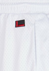Hvite idrettsshorts laget av pustende materiale, med små hullmønstre, elastisk midjebånd og en svart og rød brodert logo.
