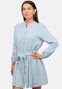 Abito blu chiaro con bottoni realizzato in tessuto testurizzato, caratterizzato da un colletto alla cinese e una vita legata. Maniche lunghe con polsini arricciati.