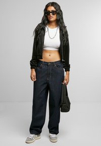 Svart fløyelsjakke med glidelås, hvit cropped topp, mørkeblå vide jeans, grå sneakers, svart veske, langt bølgete hår, oversized solbriller.
